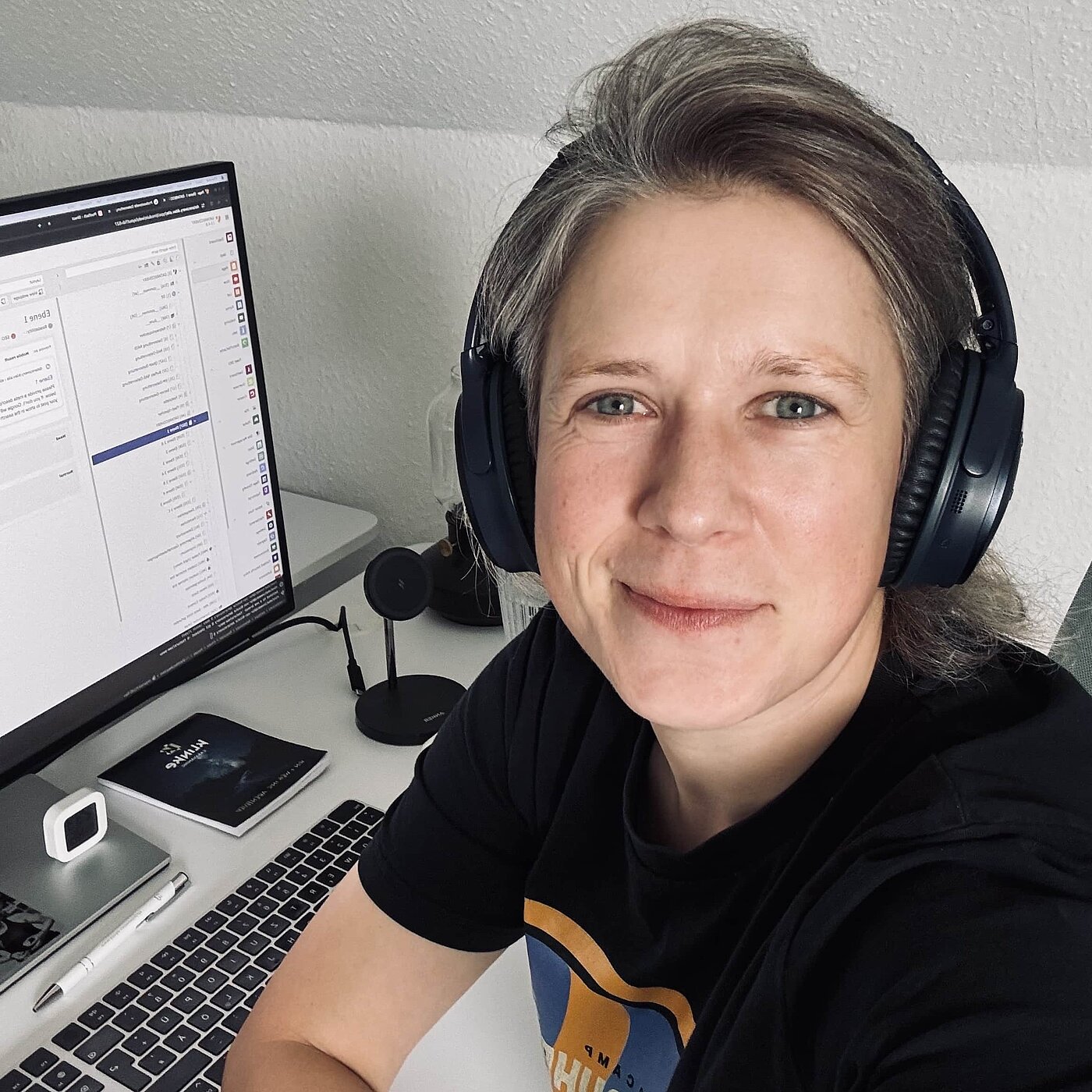 Nicole Kalinowski sitzt mit Kopfhörern an einem Schreibtisch und lächelt in die Kamera. Vor ihr steht ein Computer mit sichtbarem Bildschirminhalt. Sie trägt ein schwarzes T-Shirt und hat graue Haare. Der Arbeitsbereich wirkt modern und gut organisiert.